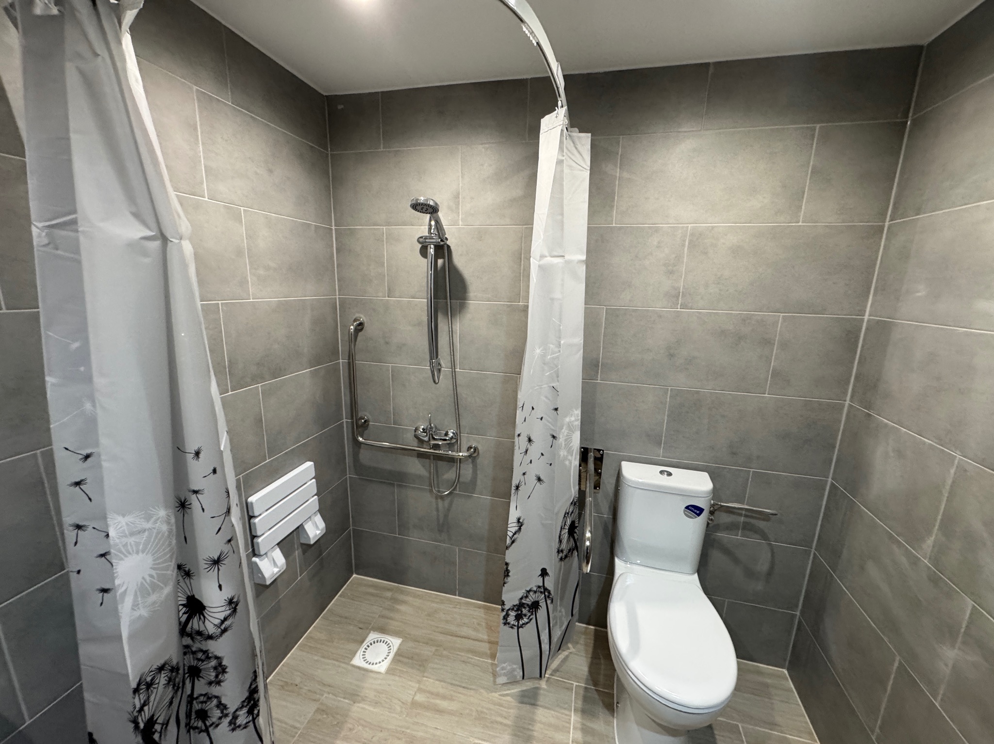 Nowoczesna łazienka w szarościach z kabiną prysznicową bez brodzika, siedziskiem i uchwytami dla niepełnosprawnych. Minimalistyczny design, płytki imitujące kamień.