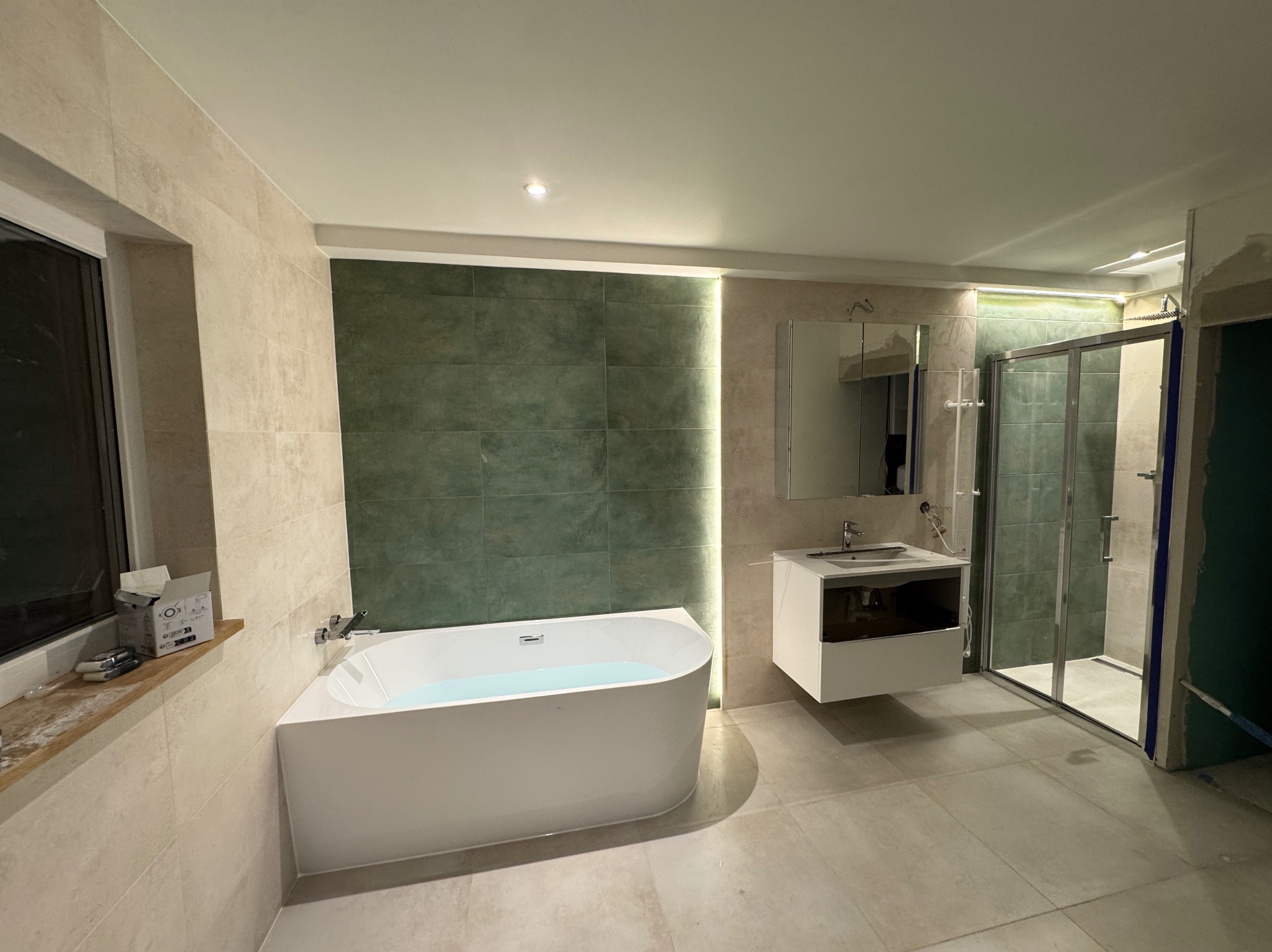 Nowoczesna łazienka z wolnostojącą wanną wypełnioną wodą, kabiną prysznicową i podwieszaną szafką z umywalką. Ściany wyłożone płytkami w odcieniach beżu i zieleni.