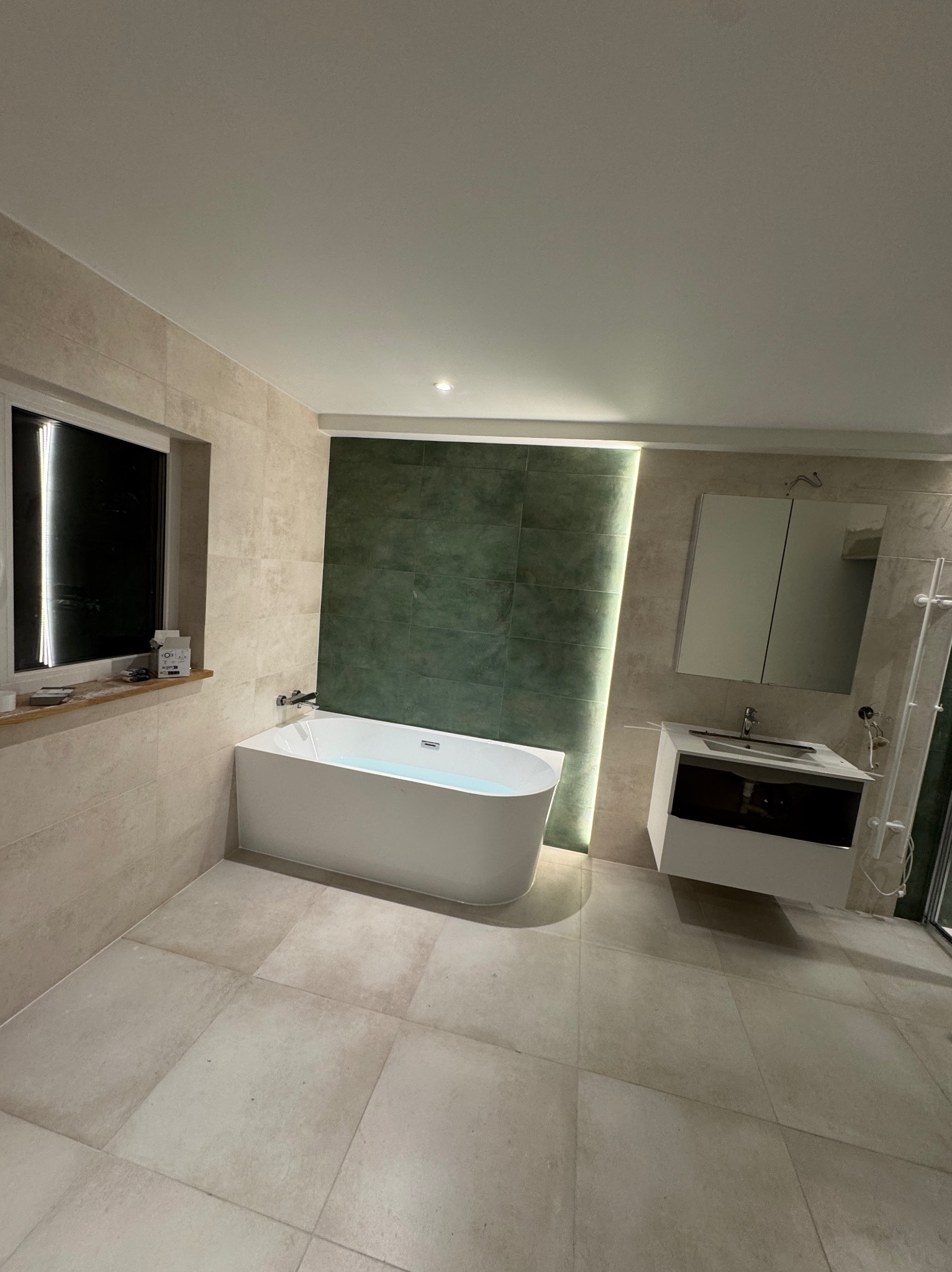 Nowoczesna łazienka z wolnostojącą wanną, beżowymi płytkami i zieloną ścianą akcentującą, oświetloną listwą LED. Minimalistyczna szafka z umywalką.