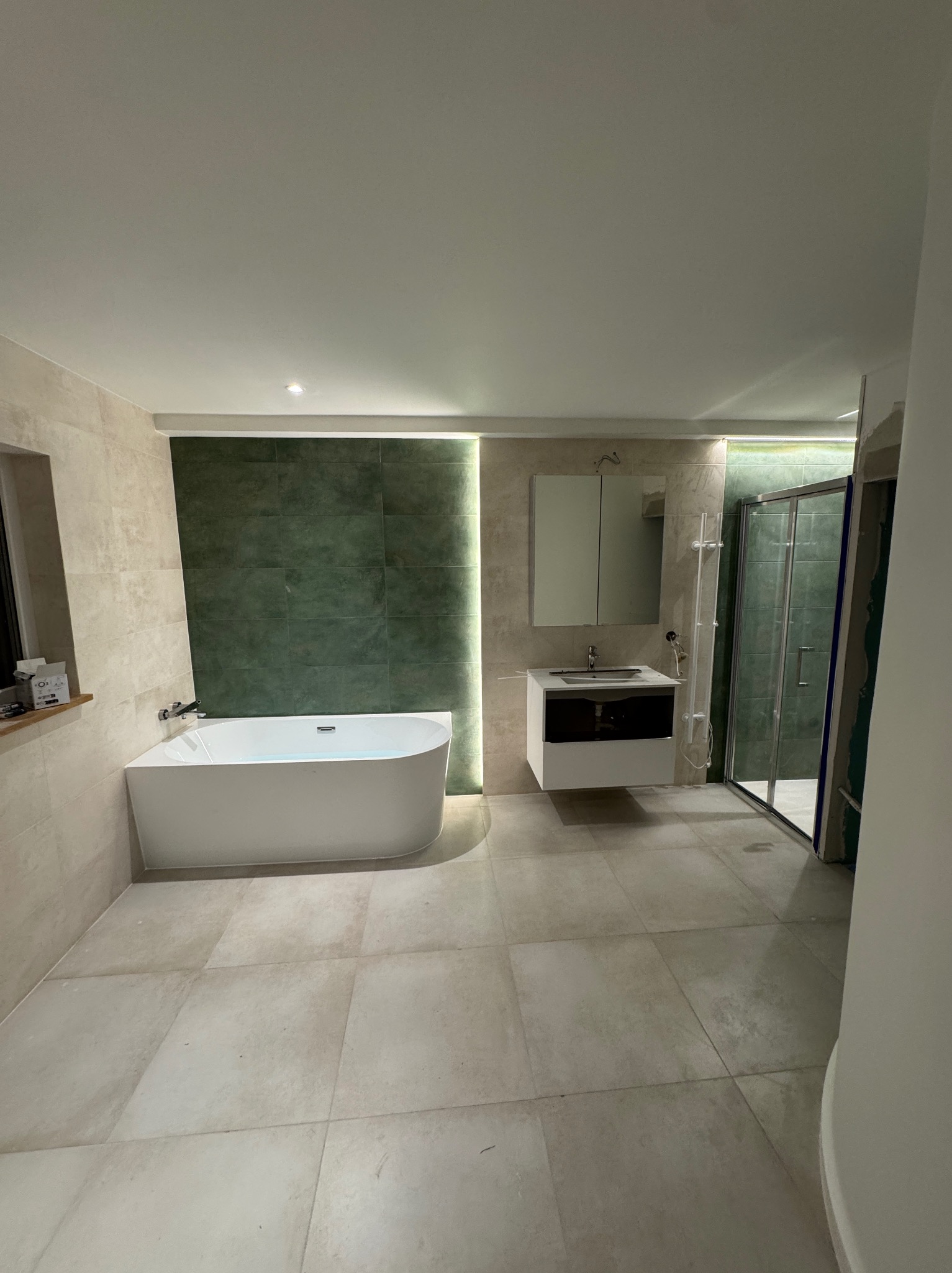 Nowoczesna łazienka z wanną, umywalką i kabiną prysznicową. Beżowe płytki podłogowe i ścienne, zielona ściana z podświetleniem. Minimalistyczny design.