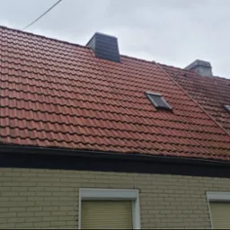 Częściowo umyty dach z czerwonej dachówki, widoczne dwa kominy, okna dachowe i antena satelitarna. Na dole fragment elewacji z numerem domu.