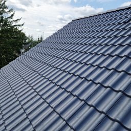 Star sp. z o.o. - Błyszczący, grafitowy dach z dachówki ceramicznej w jodełkę, widok z góry pod kątem. W tle drzewa i błękitne niebo z chmurami. Widoczny komin z cegły.