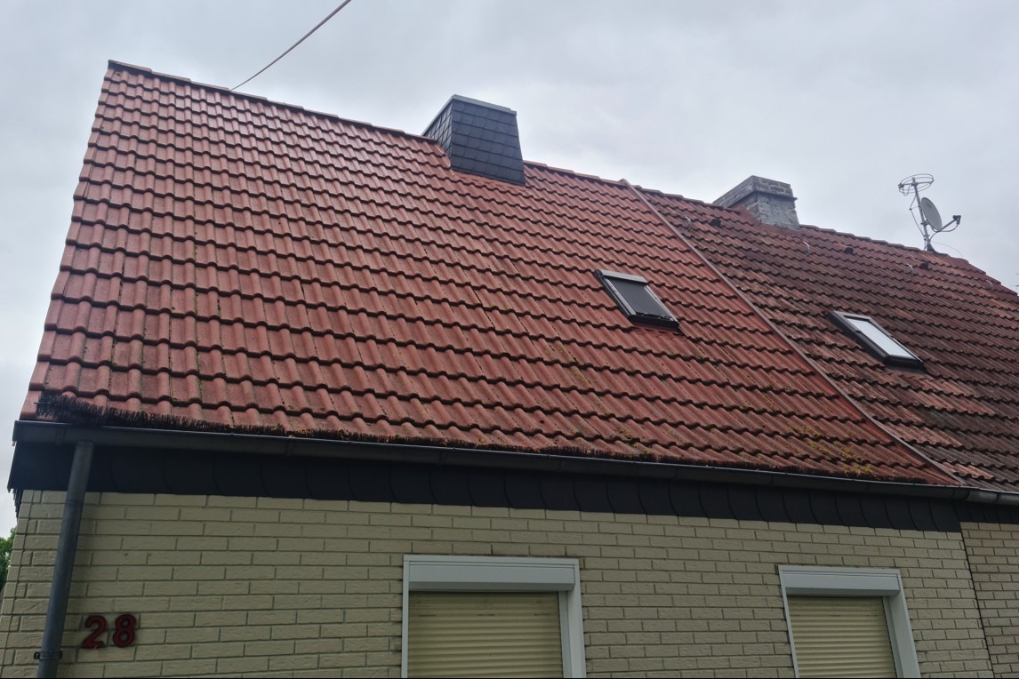 Częściowo umyty dach z czerwonej dachówki, widoczne dwa kominy, okna dachowe i antena satelitarna. Na dole fragment elewacji z numerem domu.