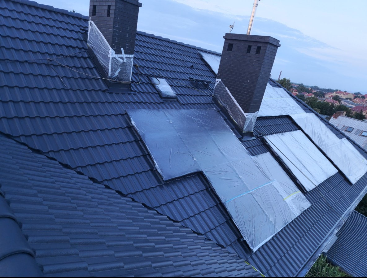 Ciemny dach z dachówki ceramicznej zabezpieczony folią przed malowaniem, widoczne kominy i fragmenty zabudowy w tle.