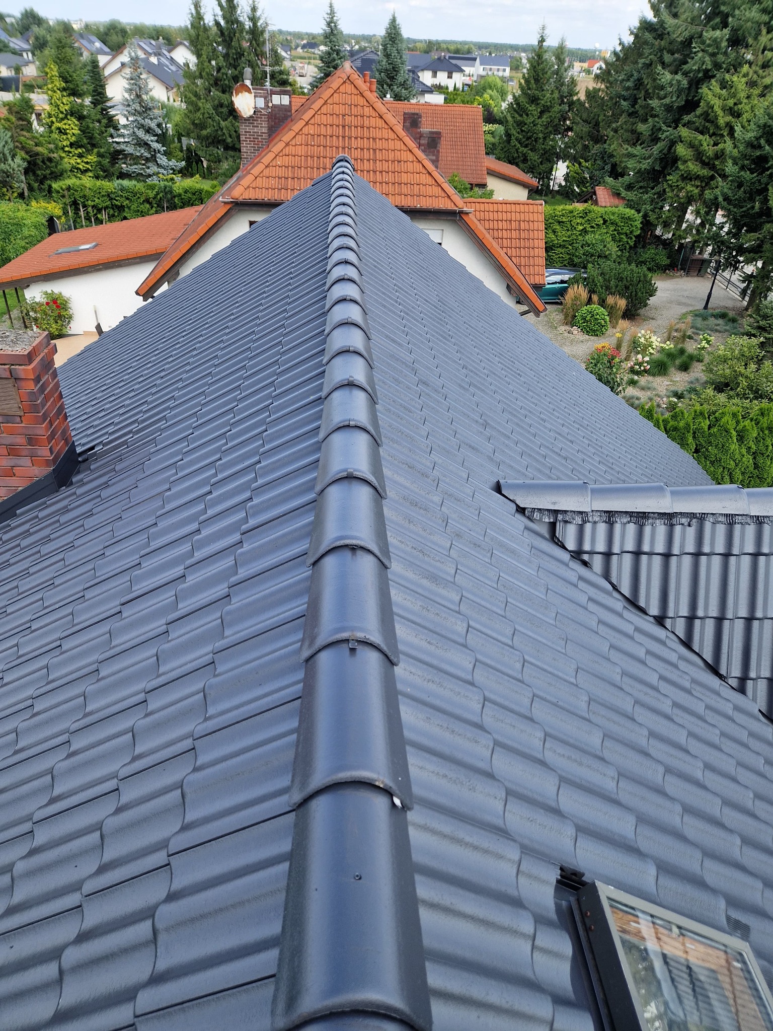 Szary, ceramiczny dach z widoczną dachówką karpiówką, z kominem w tle i fragmentem okna dachowego w prawym dolnym rogu. Widok z góry na tle zabudowy.