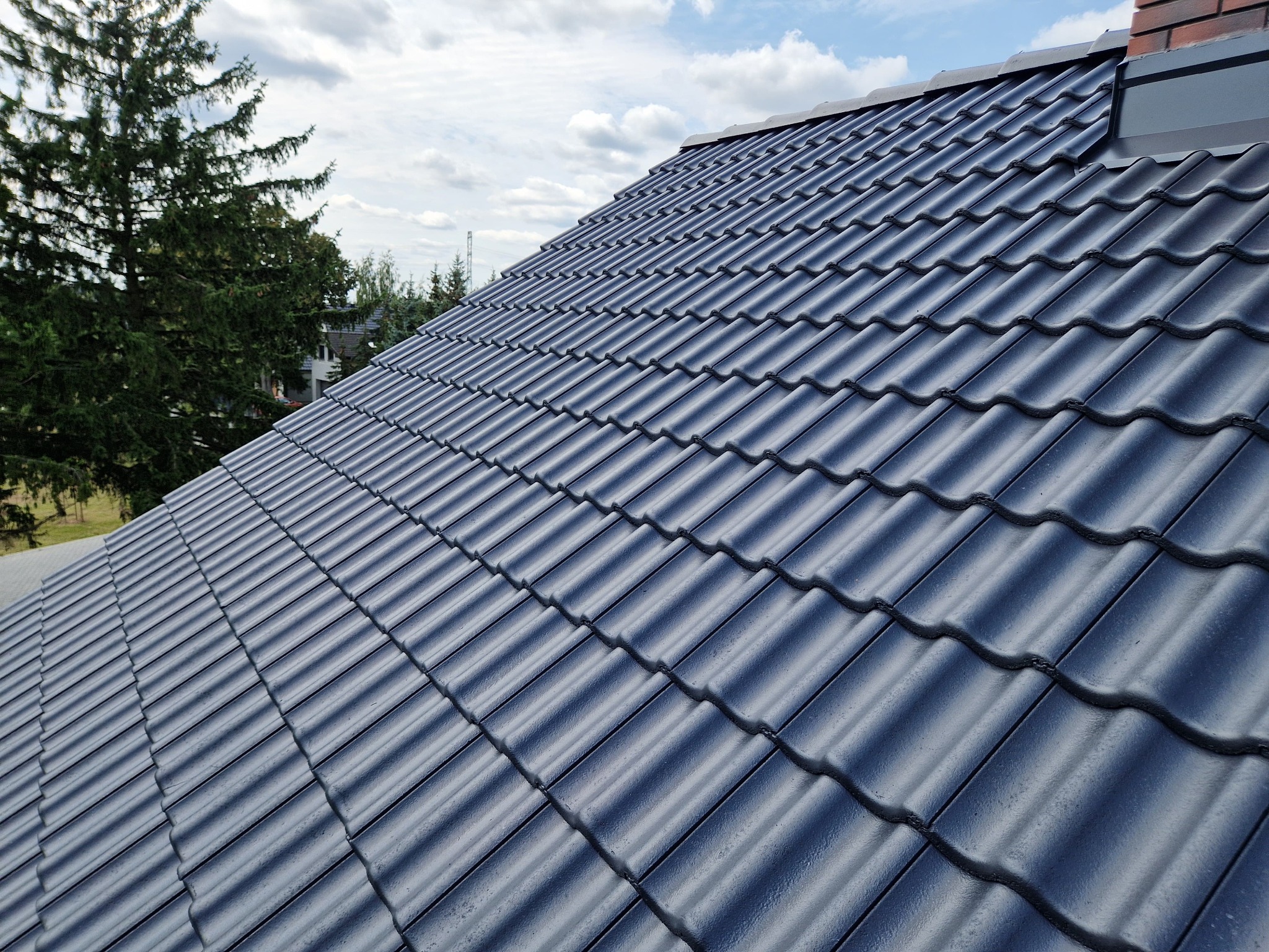 Błyszczący, grafitowy dach z dachówki ceramicznej w jodełkę, widok z góry pod kątem. W tle drzewa i błękitne niebo z chmurami. Widoczny komin z cegły.