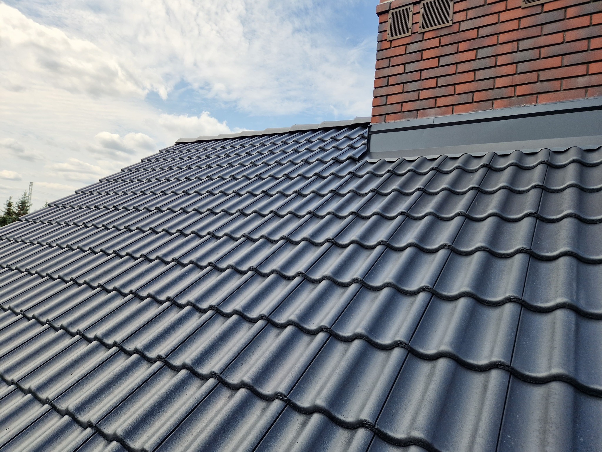 Ciemnoszara dachówka ceramiczna na skośnym dachu domu z ceglanym kominem na tle błękitnego nieba z chmurami. Widok z dołu.