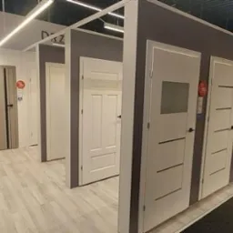 Ekspozycja różnorodnych drzwi wewnętrznych w salonie: białe modele z przeszkleniami i panelami, szare drzwi z czarnymi akcentami, jasna podłoga, sufit z oświetleniem liniowym.