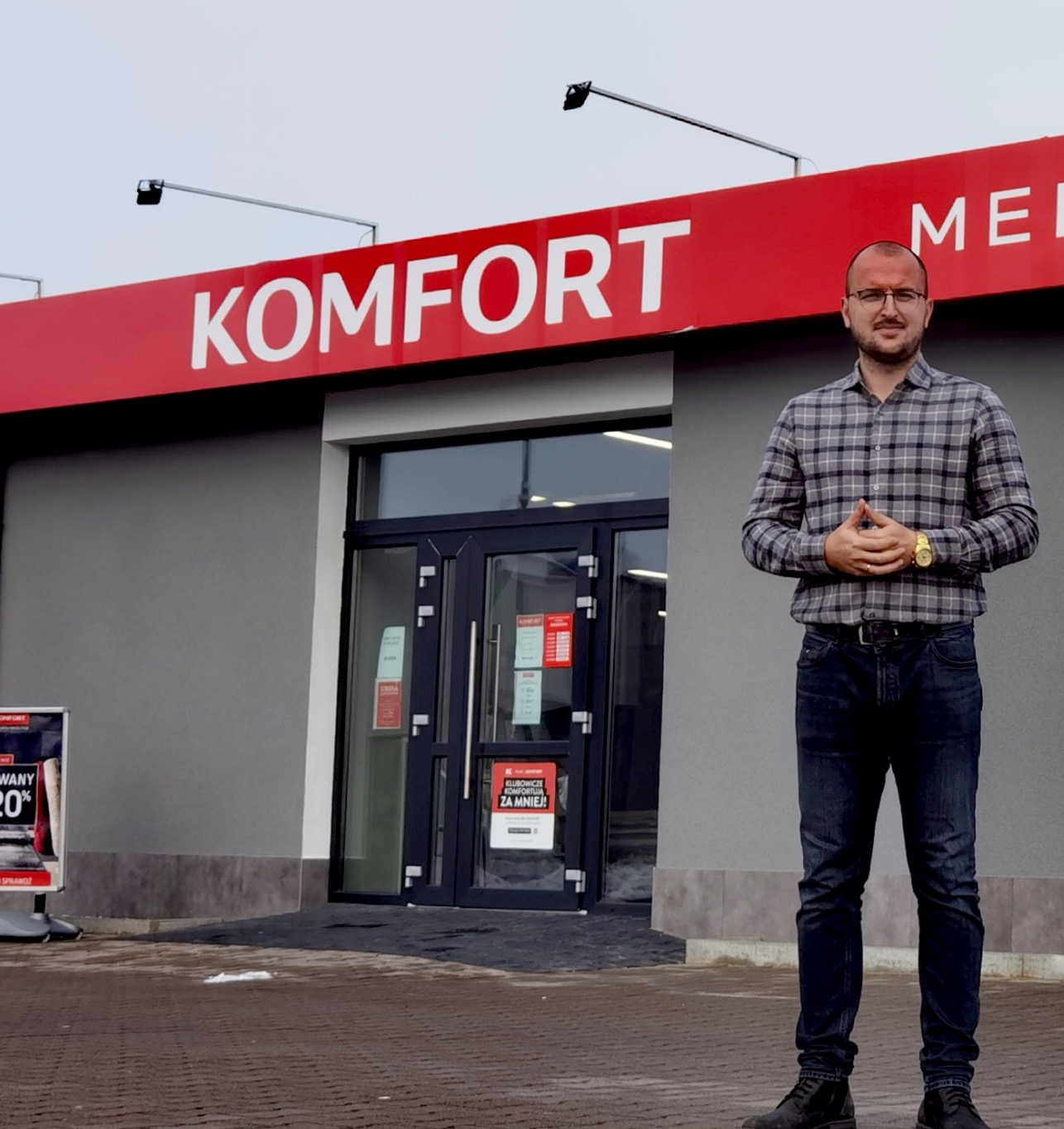Mężczyzna w okularach stoi przed wejściem do sklepu Komfort z przeszklonymi drzwiami i czerwonym szyldem. Widoczny plakat reklamowy.
