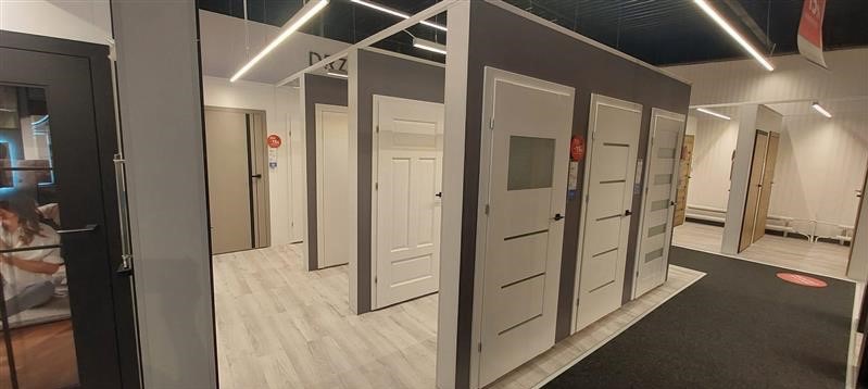 Ekspozycja różnorodnych drzwi wewnętrznych w salonie: białe modele z przeszkleniami i panelami, szare drzwi z czarnymi akcentami, jasna podłoga, sufit z oświetleniem liniowym.