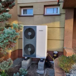 Biała pompa ciepła Vaillant, z dwoma wentylatorami, zamontowana na betonowych podstawach przy ścianie budynku z dwoma oknami. Obok rośnie iglak.