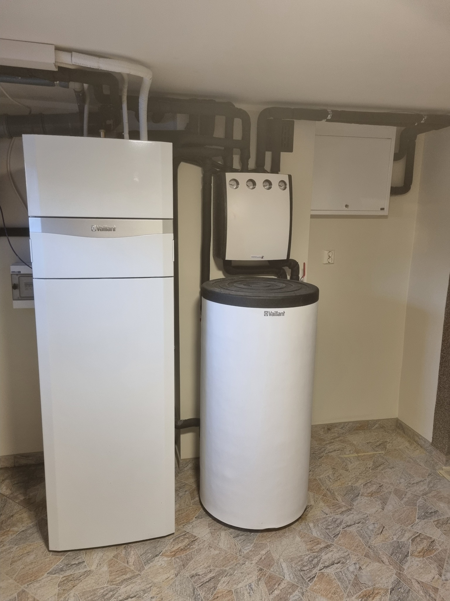 Biała pompa ciepła Vaillant i zbiornik na wodę w kotłowni z widocznymi rurami instalacyjnymi pod sufitem i brązową podłogą z płytek.