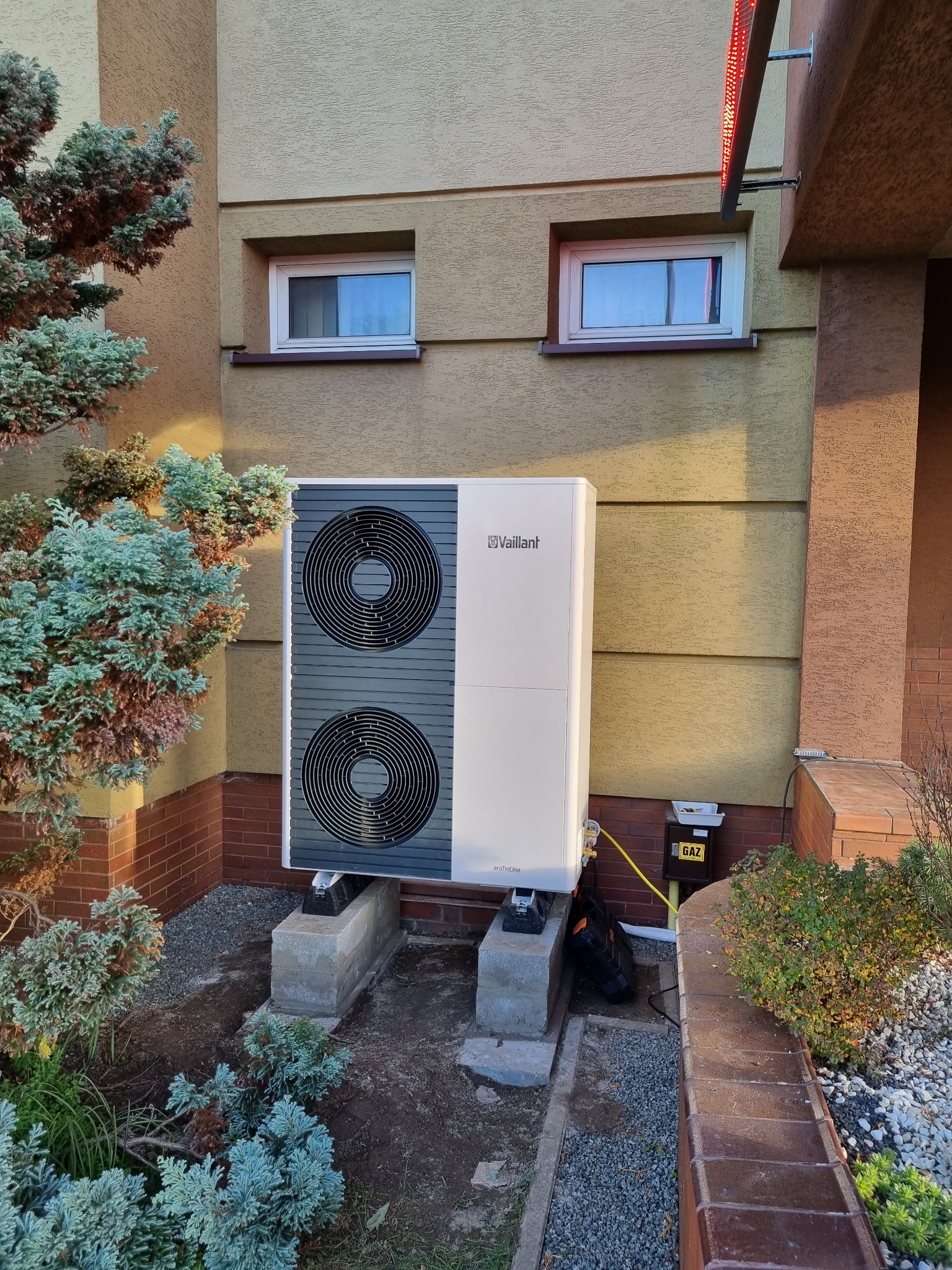 Biała pompa ciepła Vaillant, z dwoma wentylatorami, zamontowana na betonowych podstawach przy ścianie budynku z dwoma oknami. Obok rośnie iglak.