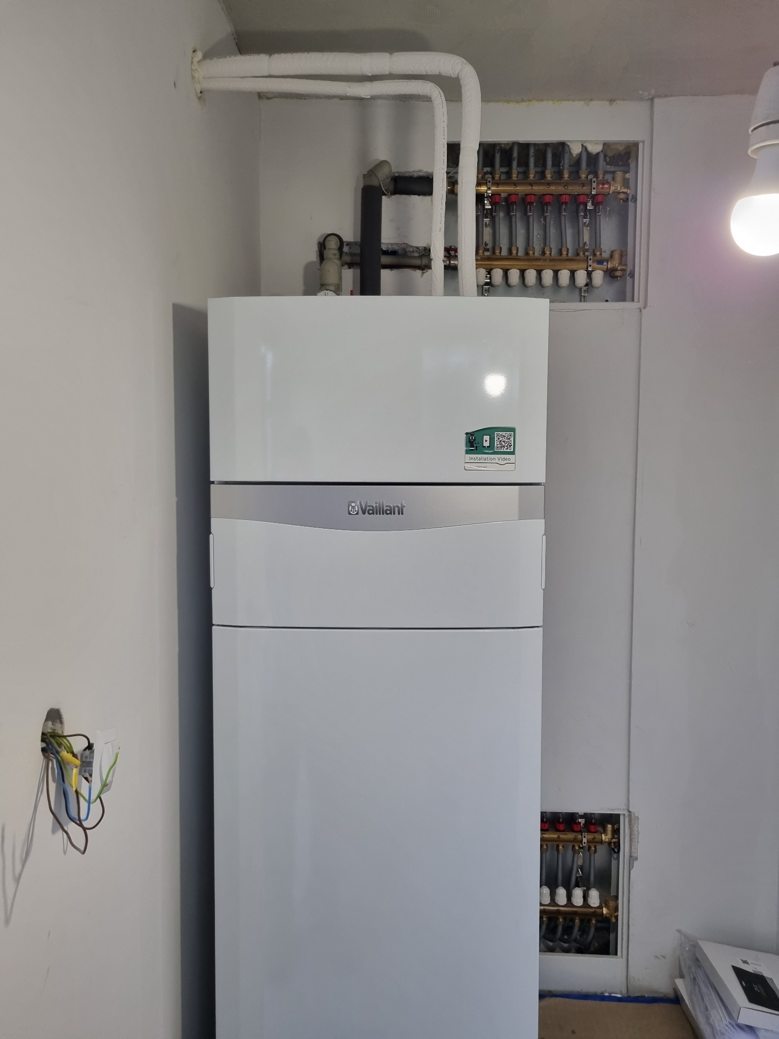 Biały, kondensacyjny piec gazowy Vaillant z rozdzielaczami ogrzewania podłogowego w ścianie. Widoczne rury i instalacja elektryczna.