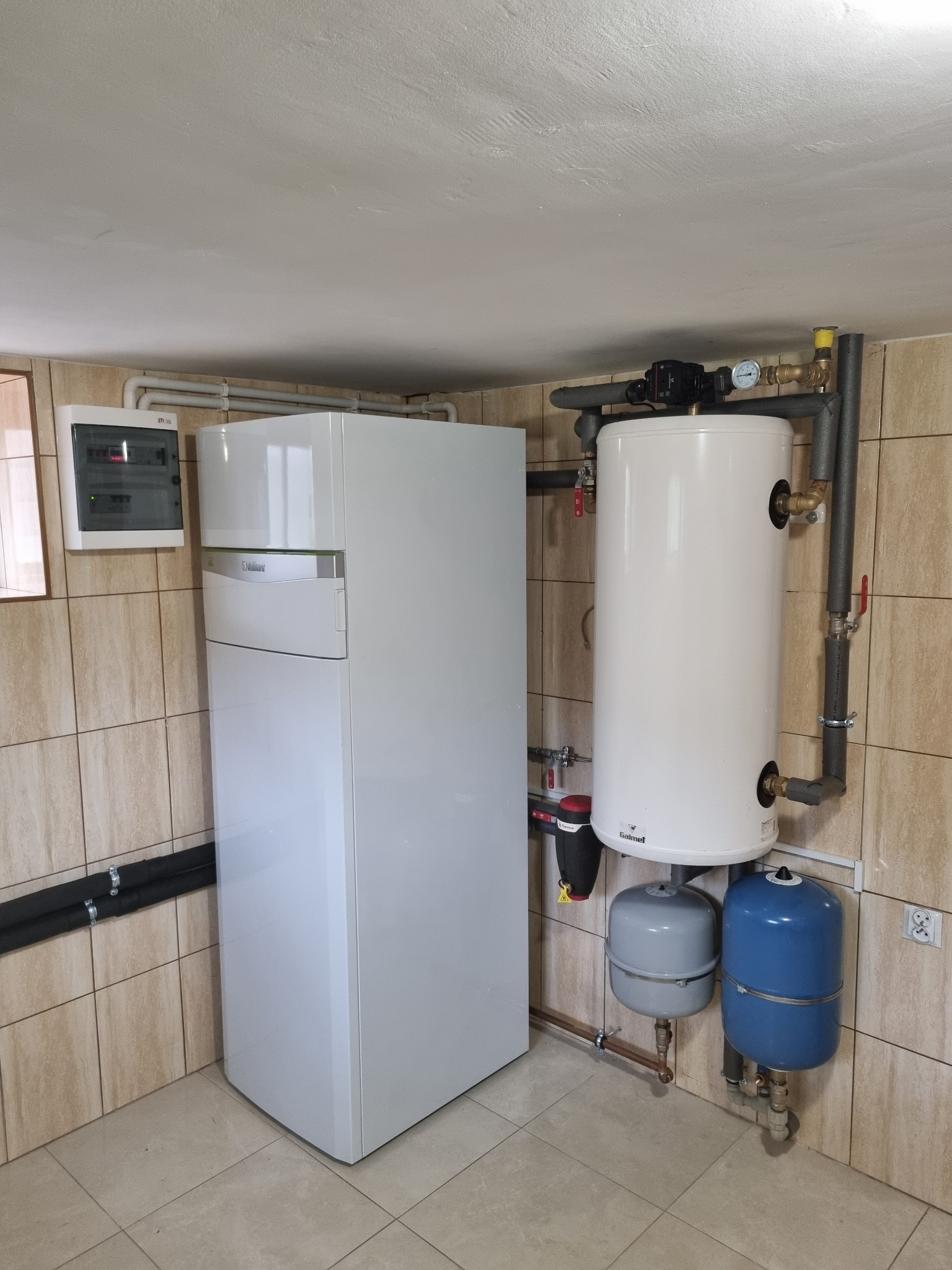 Biała pompa ciepła Vaillant z zasobnikiem Galmet i instalacją hydrauliczną w kotłowni wyłożonej beżowymi płytkami. Widoczna skrzynka elektryczna.