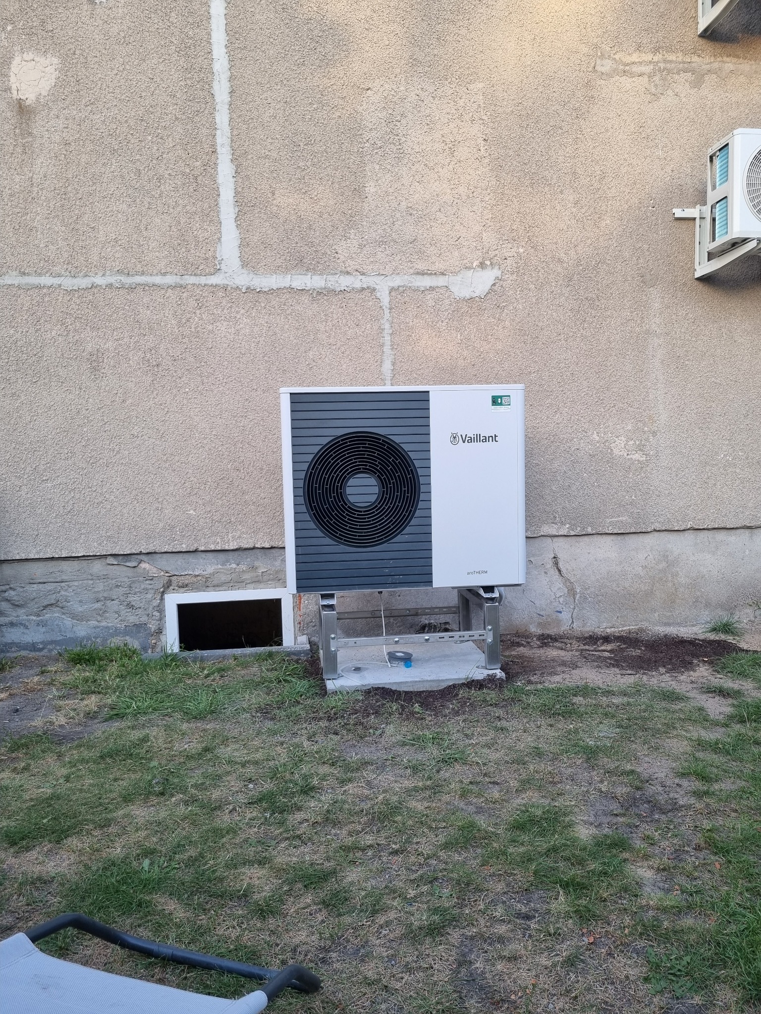 Zamontowana pompa ciepła Vaillant aroTHERM na zewnątrz budynku z widocznym innym urządzeniem klimatyzacyjnym na ścianie. Widok na trawnik.