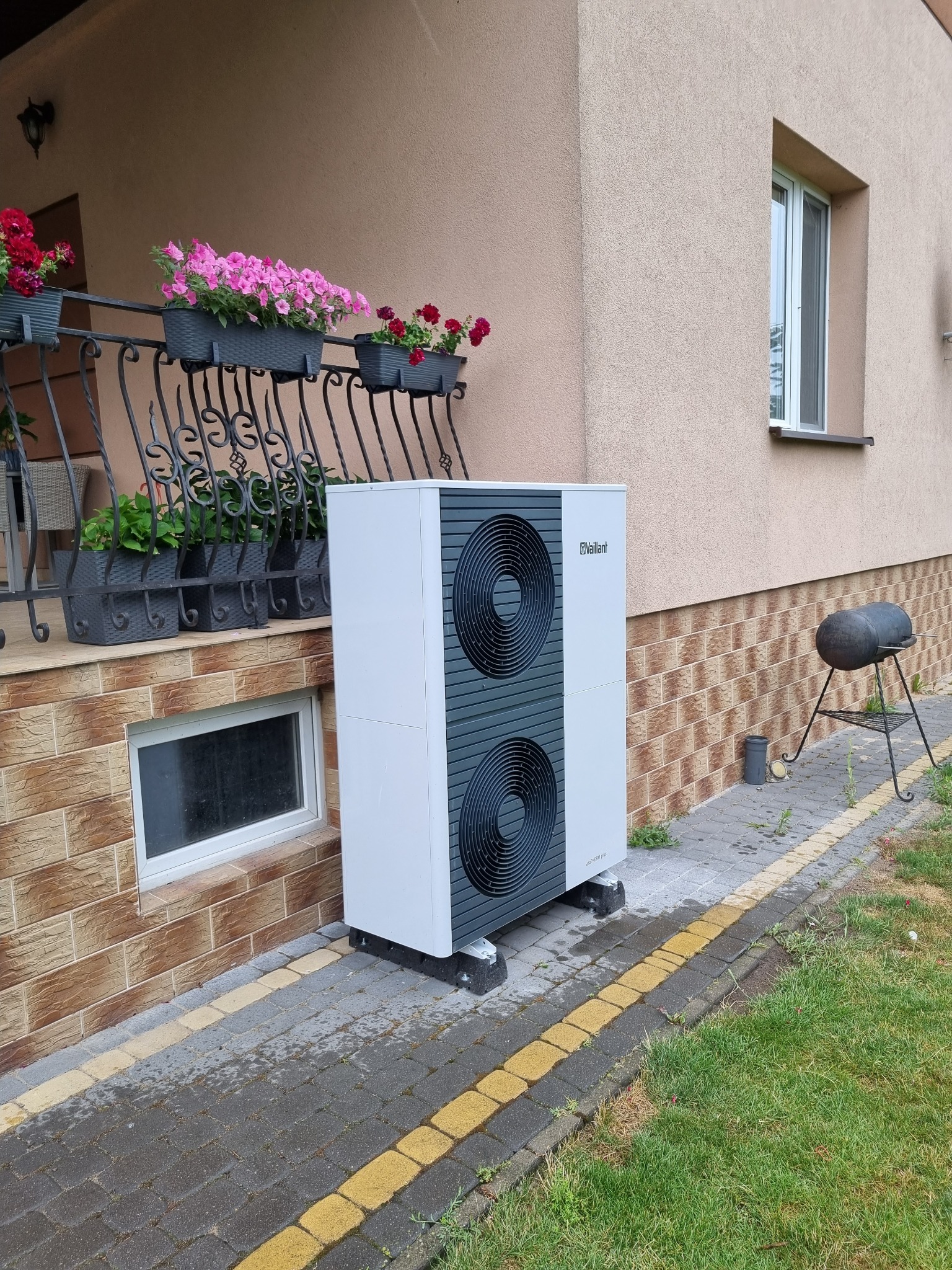 Biała pompa ciepła Vaillant zamontowana przy beżowym domu z kwiatami na balkonie. Widoczny grill na zewnątrz. Ujęcie z poziomu trawnika.