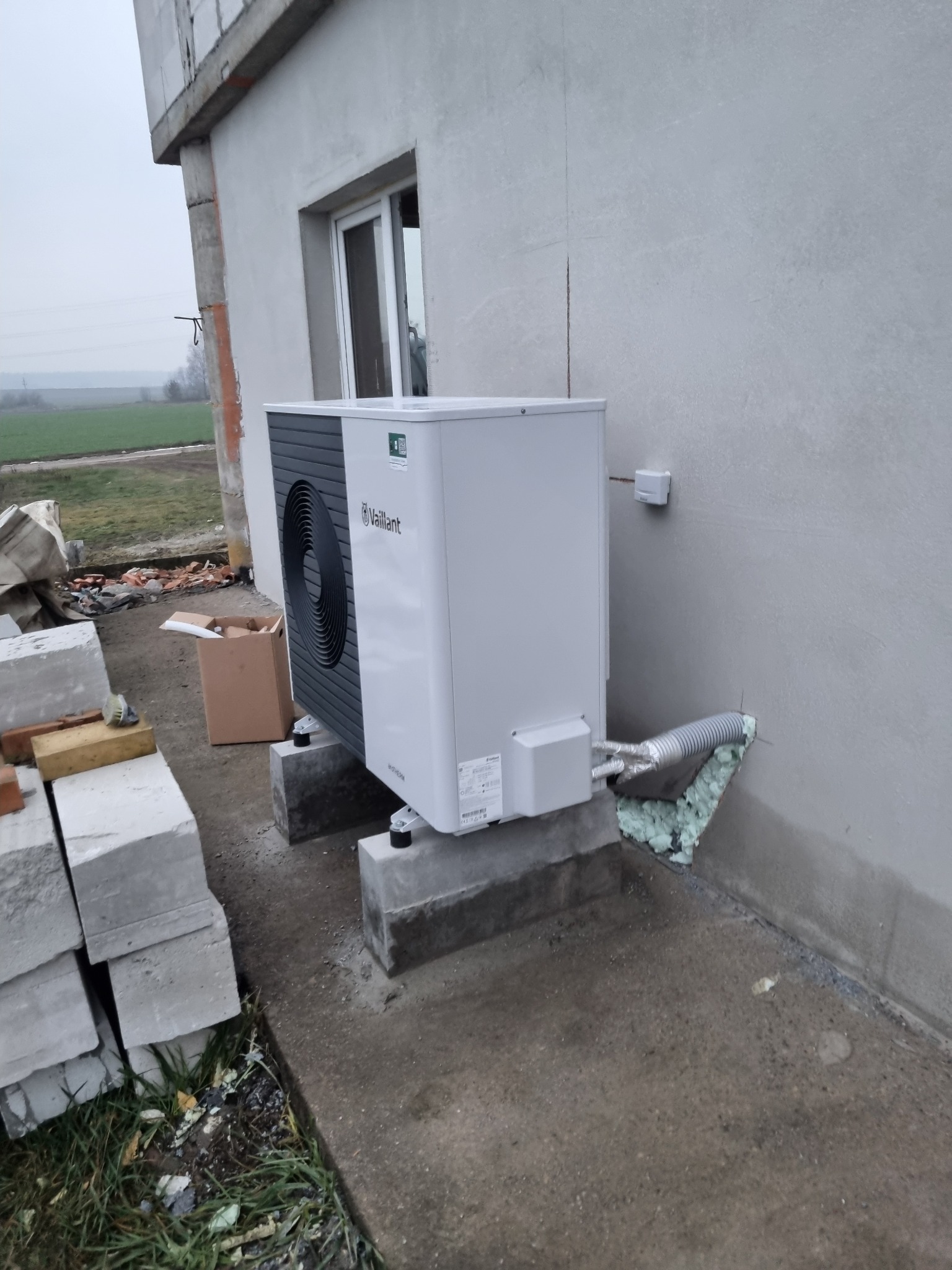Biała pompa ciepła Vaillant zamontowana na betonowych podstawach przy ścianie budynku w trakcie wykańczania. Widoczne przewody i materiały izolacyjne.