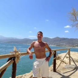 Uśmiechnięty mężczyzna oparty o drewnianą barierkę na tle morza i gór. Widoczny tatuaż na nodze, krótki naszyjnik. Słoneczny dzień.