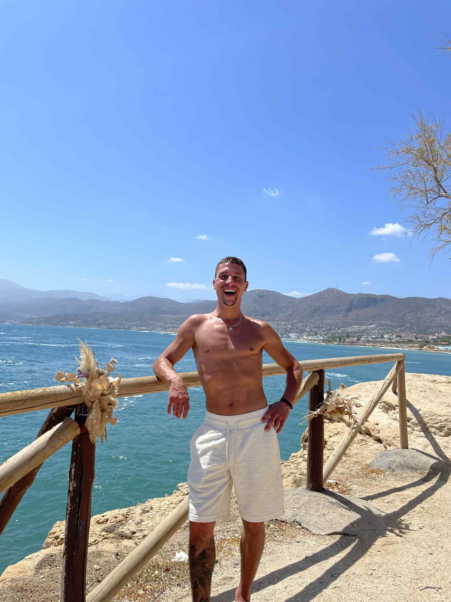 Uśmiechnięty mężczyzna oparty o drewnianą barierkę na tle morza i gór. Widoczny tatuaż na nodze, krótki naszyjnik. Słoneczny dzień.