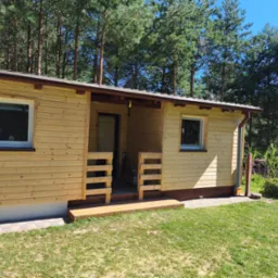 Drewniany domek letniskowy z jasną elewacją, widoczne okna z białymi ramami, zadaszone wejście. W tle wysokie drzewa iglaste i błękitne niebo. Ujęcie w słoneczny dzień.