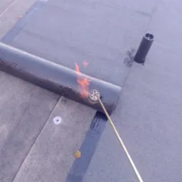 Prace dekarskie: palnik dekarski w akcji podczas zgrzewania papy na dachu. Widoczna spoina, fragment rury wentylacyjnej i pojedynczy liść na powierzchni dachu.