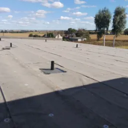 Szary dach płaski z widocznymi kominkami wentylacyjnymi i elementami mocującymi na tle błękitnego nieba z chmurami i wiejskiego krajobrazu z drzewami.