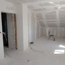 Poddasze w trakcie prac wykończeniowych: ściany i sufit pokryte płytami karton-gips, widoczne narzędzia, krzesło i przewody elektryczne. Stan surowy przed malowaniem.