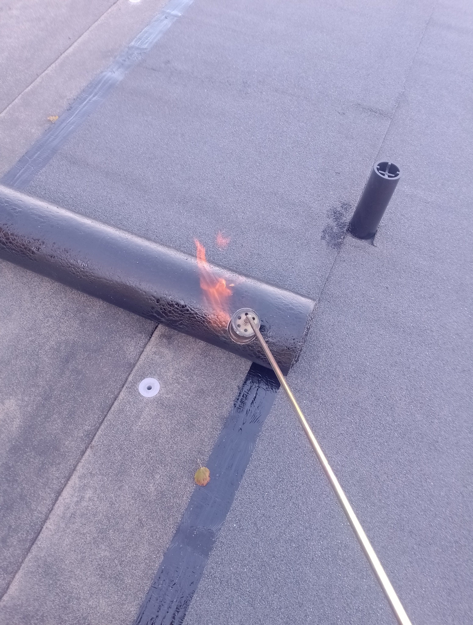 Prace dekarskie: palnik dekarski w akcji podczas zgrzewania papy na dachu. Widoczna spoina, fragment rury wentylacyjnej i pojedynczy liść na powierzchni dachu.