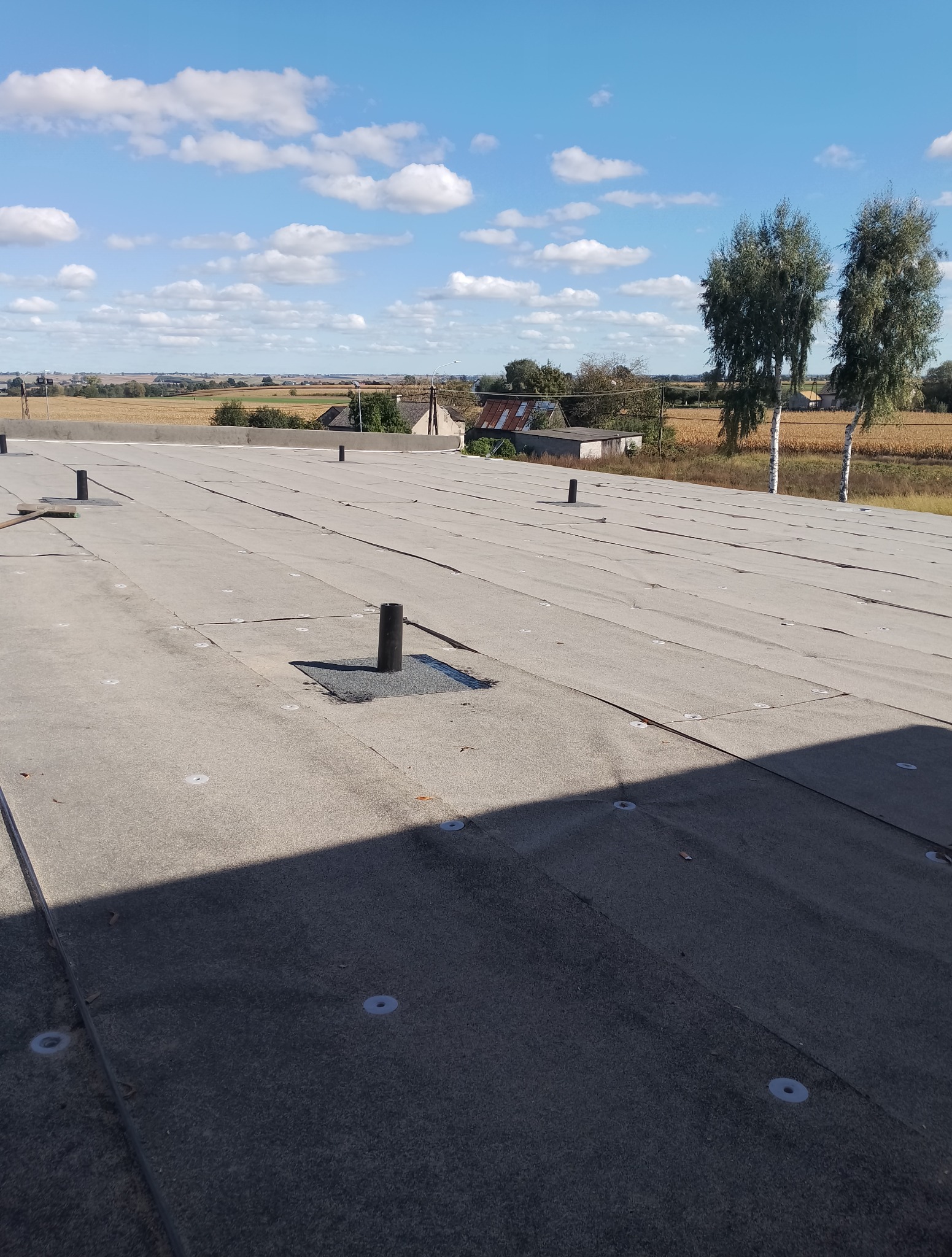 Szary dach płaski z widocznymi kominkami wentylacyjnymi i elementami mocującymi na tle błękitnego nieba z chmurami i wiejskiego krajobrazu z drzewami.