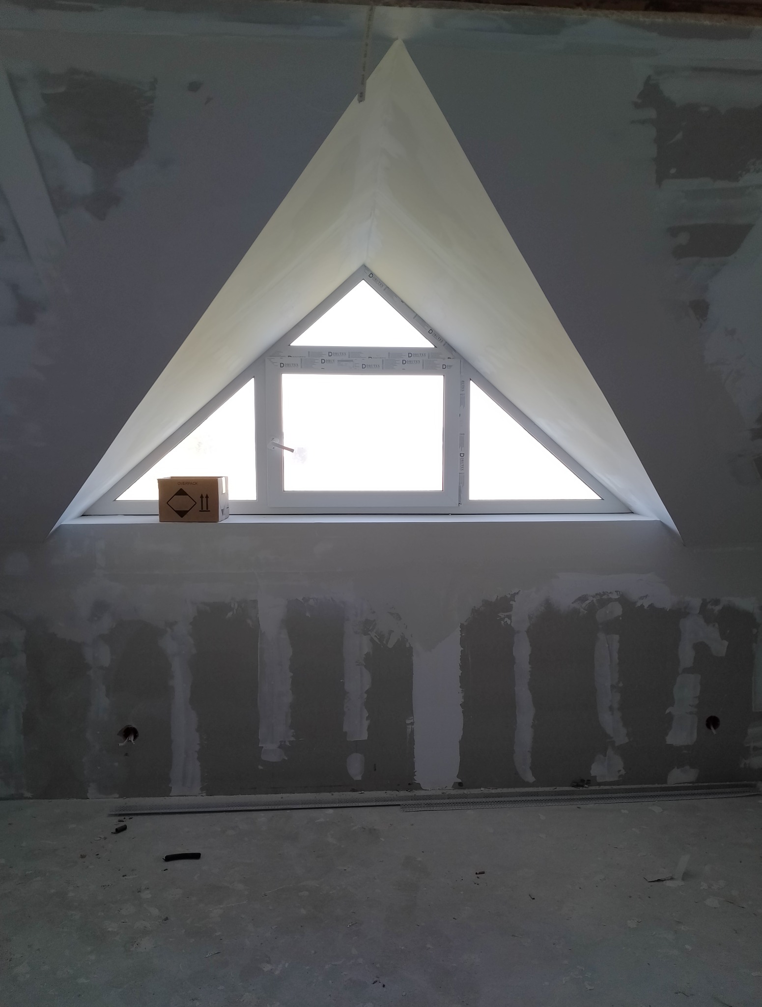 Poddasze w trakcie wykończenia: trójkątne okno, ściany z płyt karton-gips, częściowo zaszpachlowane. Na parapecie stoi karton. Widoczne ślady po instalacjach.