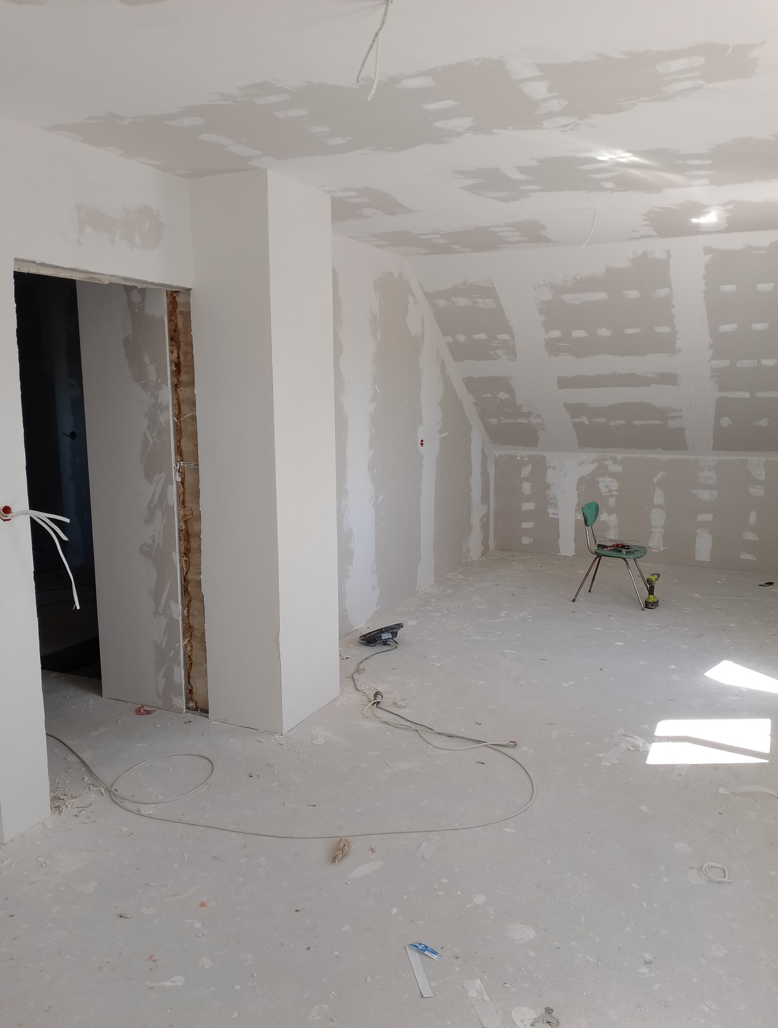 Poddasze w trakcie prac wykończeniowych: ściany i sufit pokryte płytami karton-gips, widoczne narzędzia, krzesło i przewody elektryczne. Stan surowy przed malowaniem.