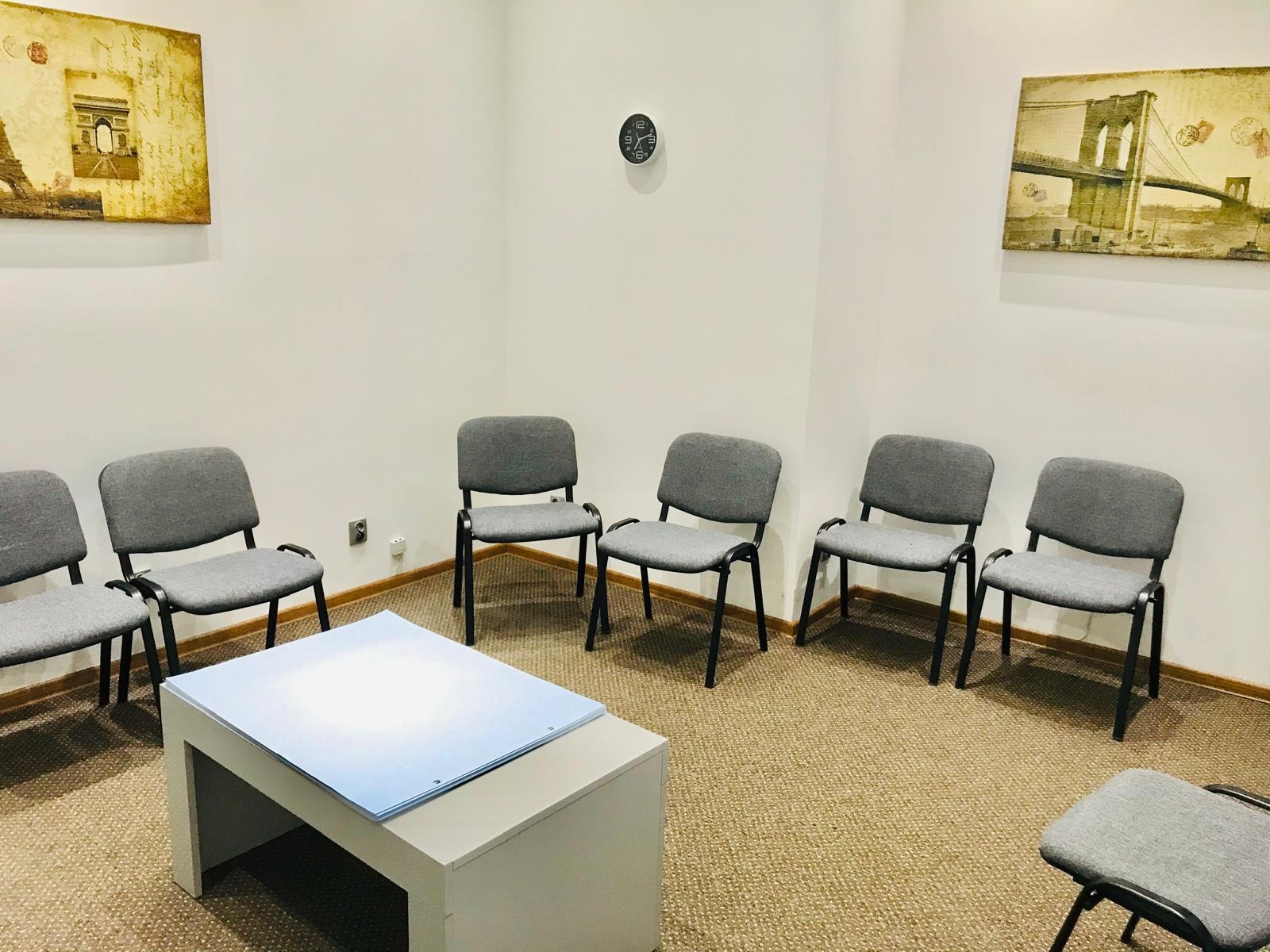 Jasne wnętrze poczekalni: krzesła w szarym obiciu ustawione wokół niskiego stolika z niebieskimi kartkami. Na ścianach obrazy z motywami miejskimi i zegar.