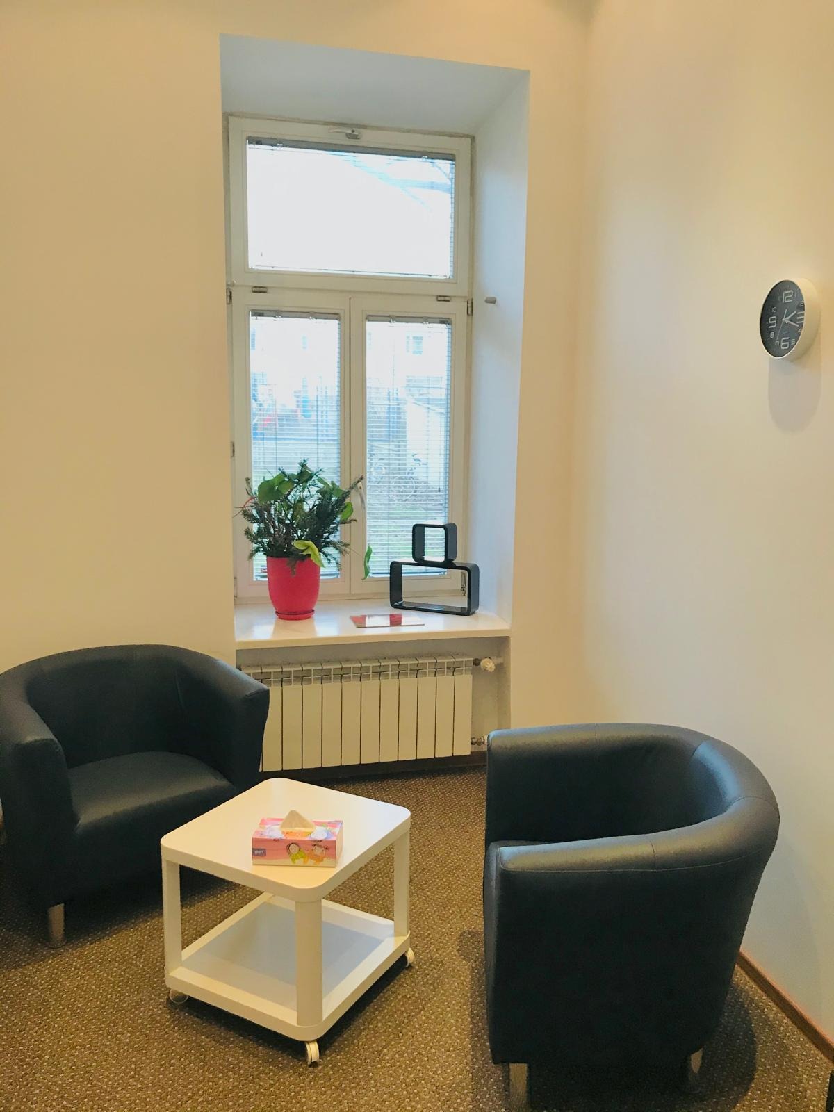 Jasne wnętrze gabinetu z dwoma czarnymi fotelami, białym stolikiem z chusteczkami i oknem z rośliną w doniczce. Na ścianie okrągły zegar.