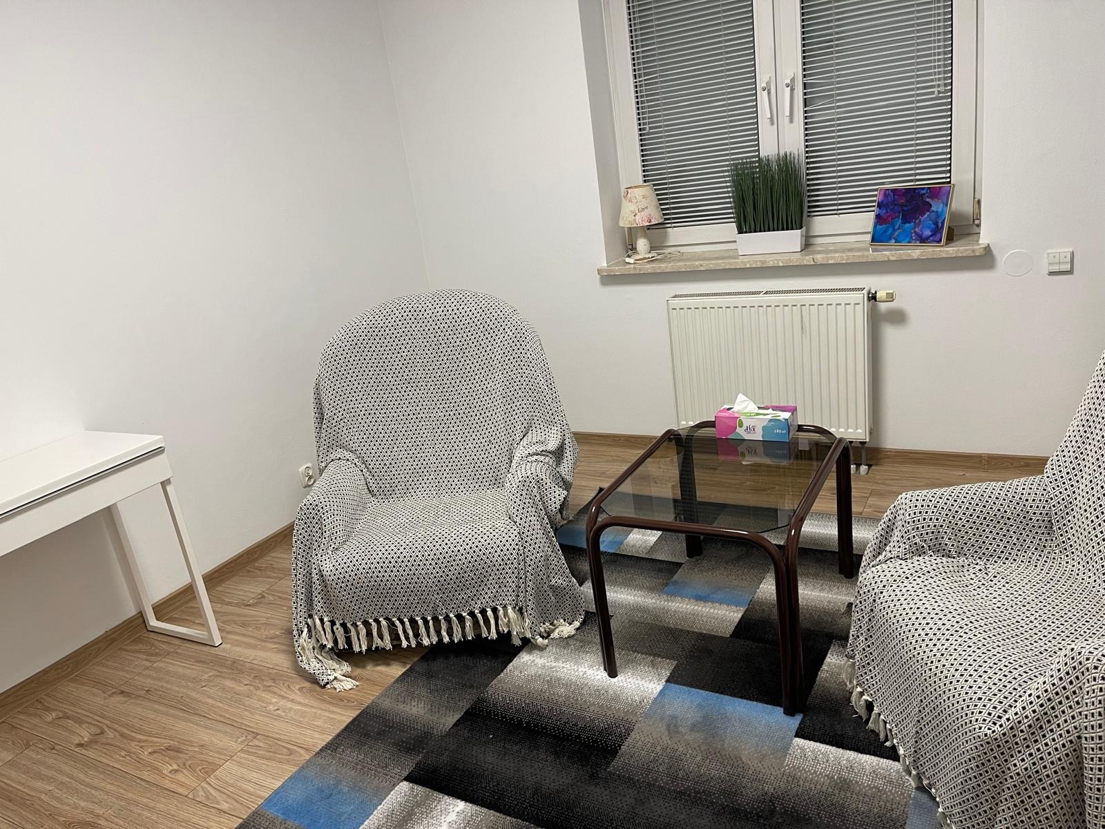 Pokój z dwoma fotelami w pokrowcach, stolikiem i dywanem w geometryczne wzory. Na parapecie lampka, roślina i obrazek. Neutralne, jasne wnętrze.