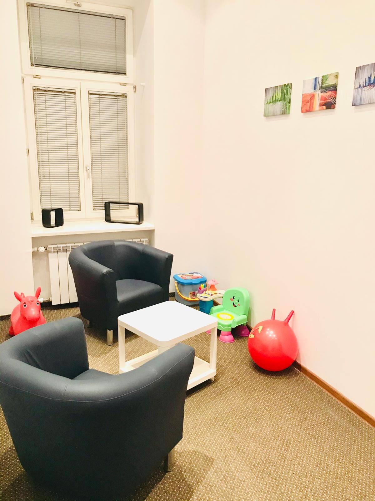 Pokój z dwoma fotelami, stolikiem i zabawkami dla dzieci, przy oknie grzejnik, a na ścianie obrazy. Neutralne kolory ścian i dywanu tworzą spokojną atmosferę.