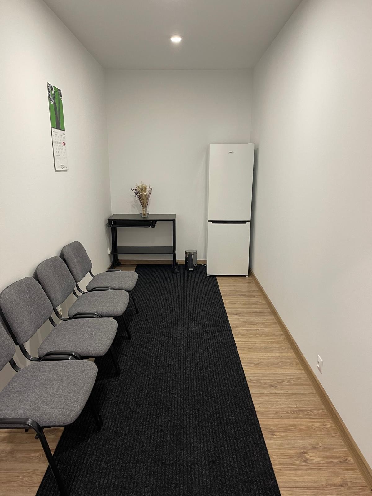 Poczekalnia: rząd szarych krzeseł, czarny dywan, stolik z suchymi kwiatami i biała lodówka. Minimalistyczne wnętrze, jasne ściany, drewniana podłoga.
