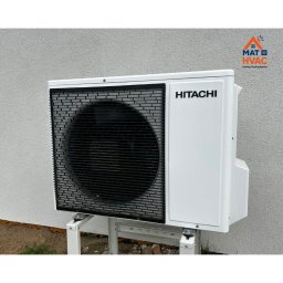 Mat HVAC - Biała zewnętrzna jednostka klimatyzacji Hitachi, zamontowana na metalowym stelażu przy jasnej ścianie budynku. Widoczne logo firmy MAT HVAC w prawym górnym rogu.