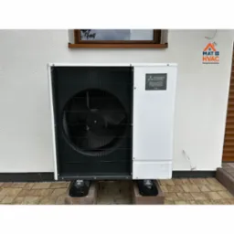 Biała pompa ciepła Mitsubishi Zubadan R32, zamontowana na zewnątrz budynku. Widoczny wentylator i logo producenta. Tło stanowi biała ściana i okno.