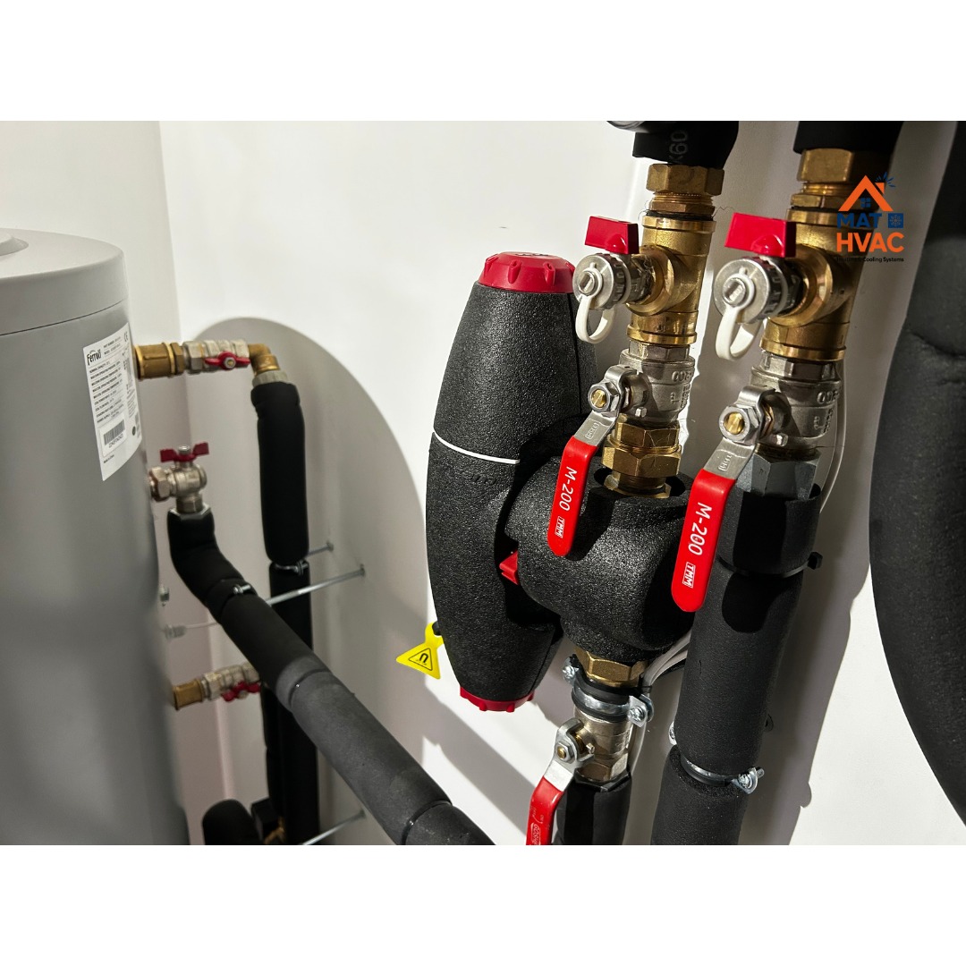 Instalacja grzewcza z zbiornikiem, rurami w izolacji i zaworami z czerwonymi dźwigniami. Elementy połączone mosiężnymi złączkami. Całość na tle białej ściany.