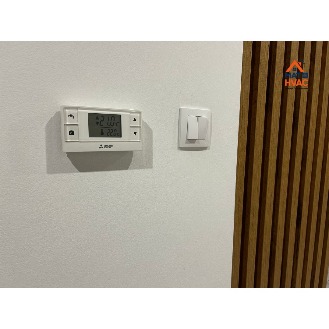 Biały panel sterowania klimatyzacji Mitsubishi Electric na ścianie, obok włącznik światła i dekoracyjne panele ścienne. Ustawiona temperatura 21°C.