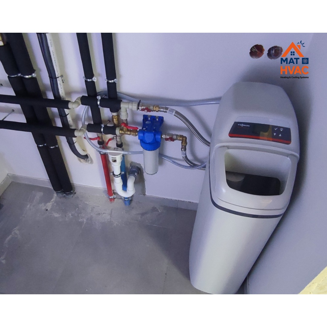 Zestaw instalacji hydraulicznej z filtrem wody i zmiękczaczem. Rury w izolacji biegną wzdłuż ściany. Widoczne złączki, zawory i elementy mocujące. Szara podłoga.