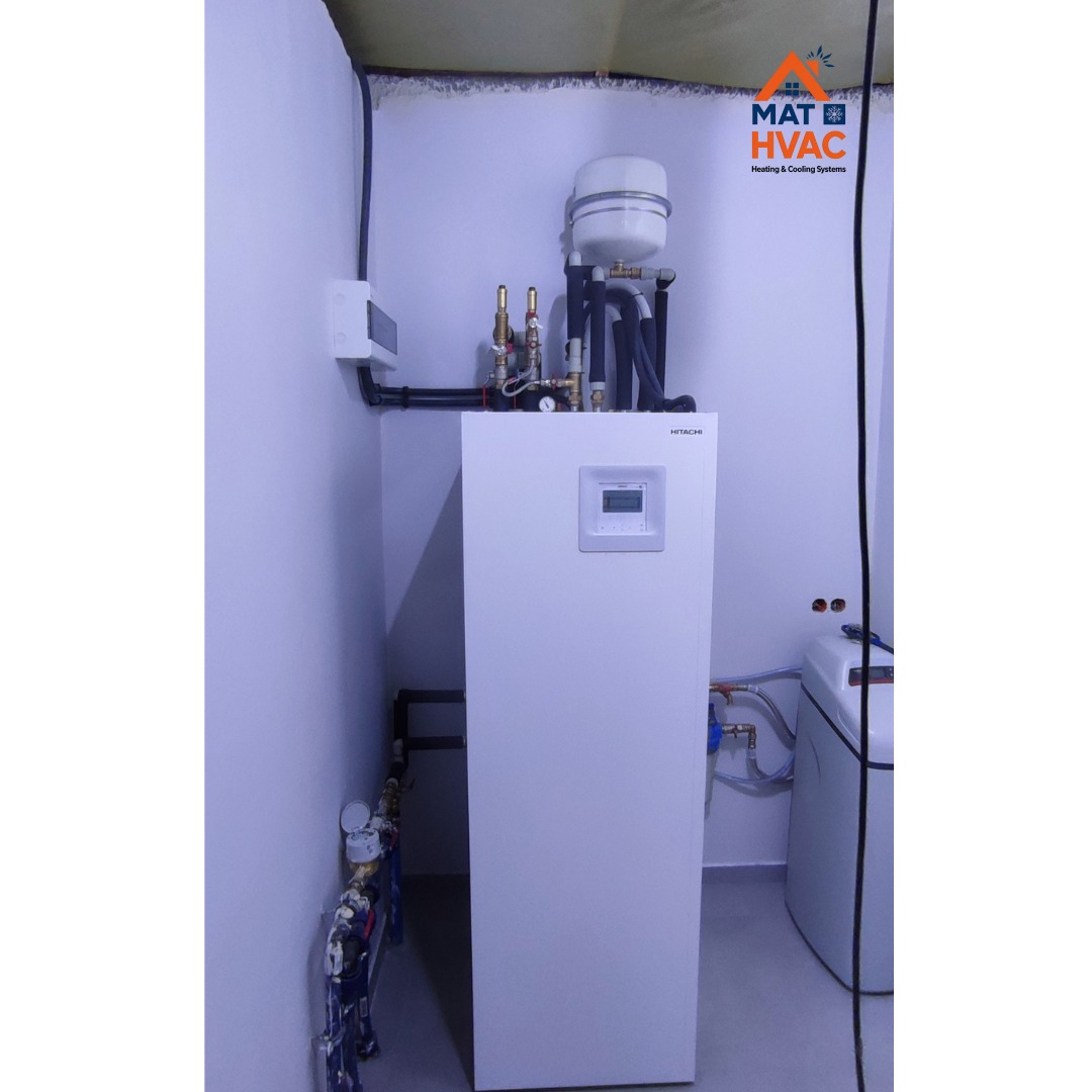 Biała pompa ciepła Hitachi w kotłowni, z widocznymi rurami i zbiornikiem wyrównawczym. Ujęcie z dołu, ukazujące instalację w narożniku pomieszczenia.