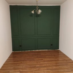 Przedsiębiorstwo Budowlane Paweł Sydlewski - Pokój po remoncie: ciemnozielona ściana z dekoracyjnymi panelami, drewniana podłoga, białe ściany boczne i sufit z lampą. Klasyczny styl wykończenia wnętrza.