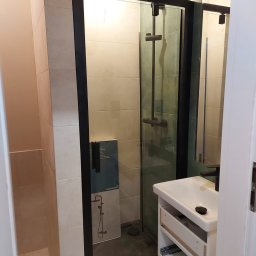 Przedsiębiorstwo Budowlane Paweł Sydlewski - Nowoczesna kabina prysznicowa z czarną ramą i deszczownicą, umywalka z szafką w trakcie montażu, jasne płytki na ścianach. Widoczny karton z grafiką baterii.