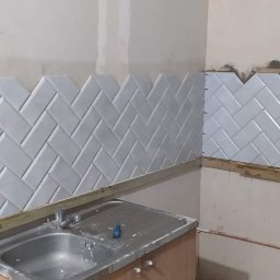 Przedsiębiorstwo Budowlane Paweł Sydlewski - Układanie białych, błyszczących płytek w jodełkę na ścianie nad zlewem w trakcie prac. Widoczne listwy pomocnicze i niedokończone krawędzie.