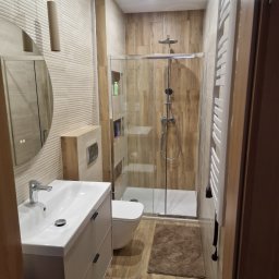Przedsiębiorstwo Budowlane Paweł Sydlewski - Jasna łazienka z kabiną prysznicową typu walk-in, umywalką z szafką i podwieszaną toaletą. Na ścianach płytki imitujące drewno, okrągłe lustro z podświetleniem.