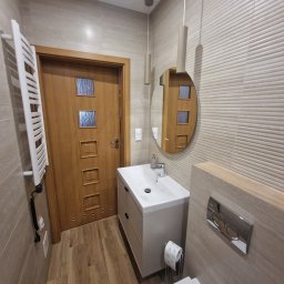 Przedsiębiorstwo Budowlane Paweł Sydlewski - Nowoczesna łazienka z beżowymi płytkami, drewnianą podłogą i drzwiami. Umywalka z szafką, okrągłe lustro i grzejnik drabinkowy. Minimalistyczne oświetlenie.