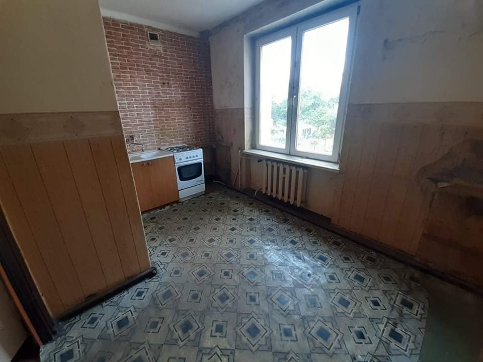 Kuchenne wnętrze przed remontem: stara podłoga w geometryczne wzory, ceglana ściana, okno z grzejnikiem i kuchenka. Widok na zaniedbane pomieszczenie.