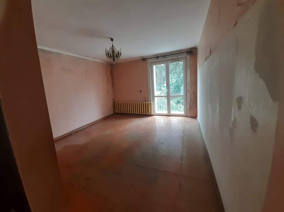 Pokój przed remontem: różowe ściany z ubytkami, stara podłoga, okno balkonowe i żyrandol. Grzejnik żeberkowy pod oknem. Widok na zieleń za oknem.